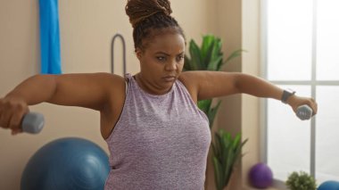 Spor salonunda dambıllarla egzersiz yapan odaklanmış bir kadın içeride azim ve zindelik gösteriyor.