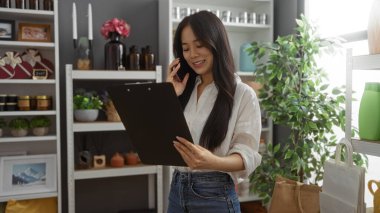 Ev dekorasyonu malzemeleriyle çevrili bir dükkanda doküman okuyan Asyalı bir kadın telefonla konuşurken, modern ve düzenli bir mağaza atmosferini vurguluyor..