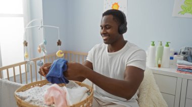 Genç adam rahat bir yatak odasında, yanında bir bebek beşiği kulaklık takmış ve gülümsüyor. İçerde huzurlu ve besleyici bir atmosfer yaratıyor..