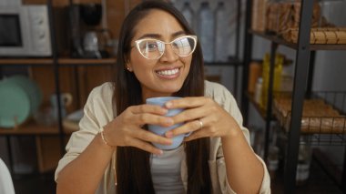 Raflar ve tabaklarla çevrili, sıcak ve davetkar bir ev atmosferi yaratan, rahat bir mutfakta elinde bardakla gülümseyen kadın..