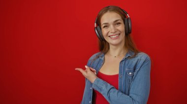Kulaklıklarla gülümseyen bir kadın canlı kırmızı duvarı işaret ediyor. Sıradan moda ve neşeli ruh hali sergiliyor..
