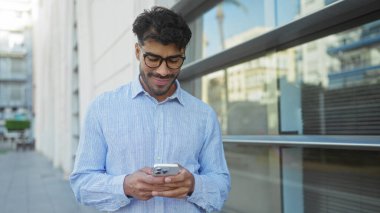 Şehir caddesinde akıllı telefona gülümseyen bir adam mimari bir arka plan ve gelişigüzel bir ortam içinde modern yaşam tarzından memnuniyet gösteriyor.