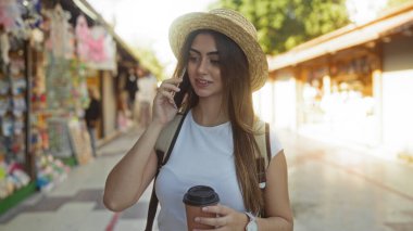 Hasır şapkalı bir kadın elinde kahveyle şehir marketi caddesinde yürüyor, telefonda sohbet ediyor, günlük açık havada geziyor, şehir yaşam tarzını aktarıyor, modern ve geleneksel unsurları harmanlıyor..