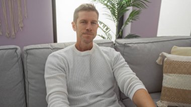 Genç adam, rahat bir oturma odasında, beyaz bir süveter giymiş, rahat ve rahat görünen, modern bir dekorasyonla, rahat bir şekilde oturuyor..