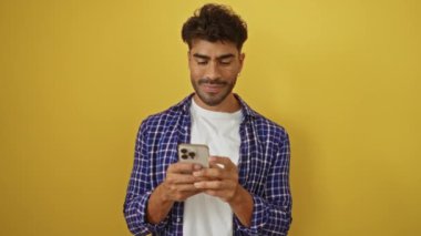 Sakallı genç adam akıllı telefonu tutuyor ve parlak sarı bir arka plana gülümserken başparmağını kaldırıyor, izole bir stüdyo ortamında pozitiflik ve güven yayıyor..