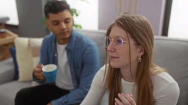 Çift rahat bir dairede kanepede oturuyor. Kahvelerini paylaşıyorlar. Kot ceketli bir adam, gözlüklü ve uzun saçlı bir kadın..