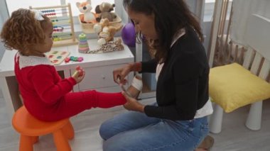 Kadın rahat bir yatak odası ortamında ayakkabılı çocuğa yardım ediyor, oyuncaklar ve beşik, aile ve aşk ön plana çıkıyor..