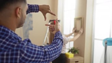 Ev dekorasyonu yapan erkek ve kadın, duvarda çerçeve tutuyorlar ve yakınlarda da takım çalışması ve ev ortamında yaratıcılık gösteriyorlar.