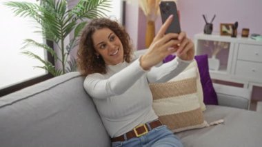 Şık oturma odasındaki kanepede selfie çeken bir kadın doğal güzelliği ve moda karışımını bitkiler ve dekorasyon geliştiren rahat bir atmosferle parlak ev ortamında gösteriyor..