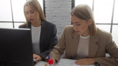 Modern bir ofis ortamında işbirliği yapan iki profesyonel kadın, belge ve bilgisayar üzerinde yoğunlaşıp, iş ortamını ön plana çıkarıyorlar..