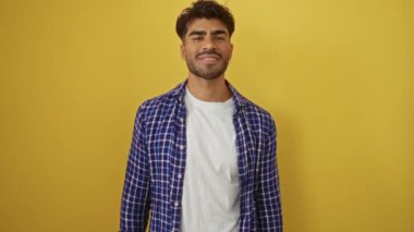 Genç adam rahat gömleğiyle canlı sarı arka plana doğru gülümsüyor. Dostane bir ifade ve davetkar bir jest sergiliyor..