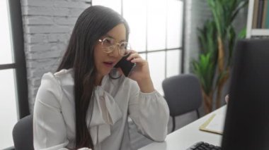 Modern ofiste telefonla konuşan, bilgisayar, gözlük ve bitkilerle görünen, odaklanmış ifade ile profesyonel Asyalı yetişkin ortamını vurgulayan bir kadın..
