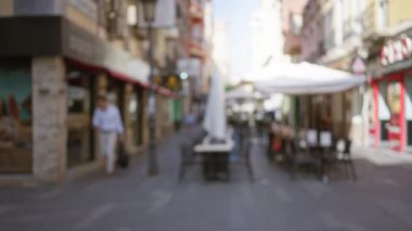 Şehir ortamında güneşli bir günde dükkanların ve açık hava yemek alanlarının önünden geçen bir adamın olduğu bulanık sokak manzarası.