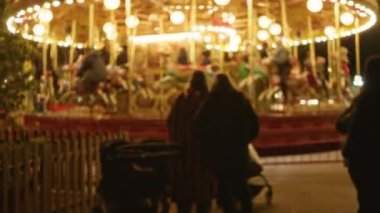 Parktaki bulanık bir atlıkarınca sahnesi. Bir grup siluetli insan ve bebek arabasıyla. Parlayan ışıklar ve bokeh efektleriyle çevrili..