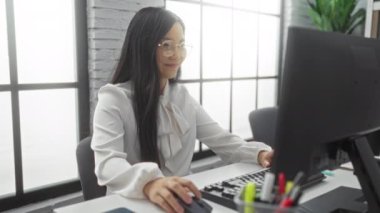 Bilgisayarda modern bir ofiste çalışan Asyalı bir kadın içerikli görünüyor ve görevleriyle meşgul..