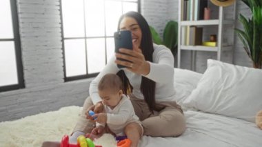 Sıcak bir yatakta bebeğiyle selfie çekerken gülümseyen bir kadın şık bir yatak odasında sevgi dolu bir aile anı sergiliyor..