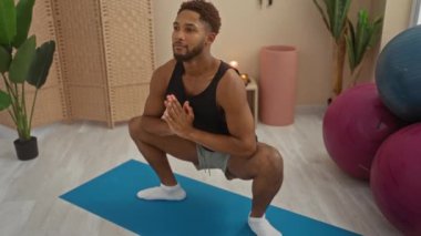 Spor salonunda yoga yapan genç bir adam siyah bir kolsuz tişört ve spor aletleri ve bitkilerle çevrili beyaz çoraplar giyiyor..