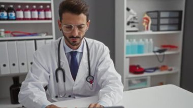 Klinikte steteskoplu laboratuvar önlüğü giyen bir adam, sağlık ekipmanlarıyla çevrili bir şekilde bilgisayar kullanırken gözlükleri ayarlıyor..