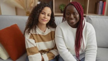 Parlak bir oturma odasında, rahat bir kanepede birlikte gülümseyen kadınlar arkadaşlığı ve sıcaklığı ön plana çıkarırlar..