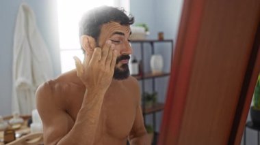 Sağlık merkezinde sakallı genç bir adam aynanın karşısına yüz kremi sürüyor.