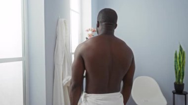 Genç adam sağIık kaplıcasında güvenle duruyor, havlu giyiyor, yumuşak ışık altında gülümsüyor, davetkar, rahat bir atmosfer yaratıyor, spa ortamının rahatlatıcı güzelliğini arttırıyor..