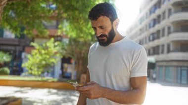 Güneşli bir günde, şehir parkında kafası karışmış bir şekilde telefonuna bakan genç İspanyol bir adam.