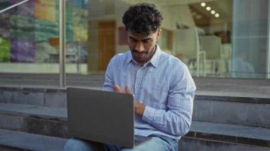 Sakallı genç adam dışarıda dizüstü bilgisayarla çalışıyor, şehir ortamında esniyor, şehirde rahat ve yakışıklı bir hava yayıyor..