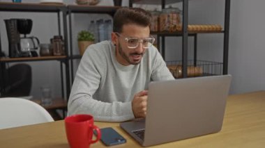 Modern ofis içinde dizüstü bilgisayarda çalışan genç adam kırmızı bardak ve ahşap masadaki akıllı telefonla arka plandaki raflar sıradan bir iş yeri ayarlamasını öneriyor..