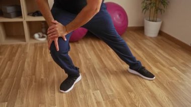 Genç bir adam spor salonunda ahşap zemini olan, mavi pantolon giyen, dizini tutan, egzersiz topları ve saksı bitkileriyle çevrili..