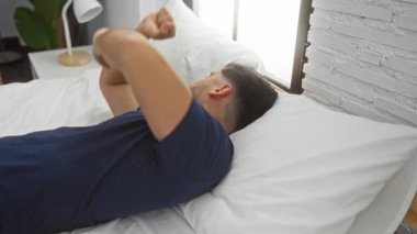 Genç adam modern bir yatak odasında yatakta dinleniyor, doğal ışıklandırma ve minimalist ev dekorasyonu elementleriyle huzurlu bir iç atmosferi yakalıyor..