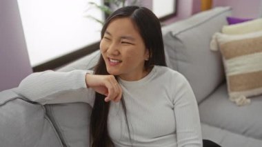 Evdeki modern bir oturma odasındaki rahat bir koltukta uzanan kadın beyaz bir kazak giyiyor ve gülümsüyor, yumuşak yastıklarla çevrili, Asya 'dan esinlenilmiş bir iç dekorda..