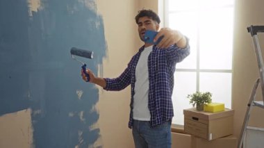 Yeni apartman dairesinde selfie çekerken duvarları silindirle boyayan genç bir adam yakınlarda kutular ve merdivenlerle kişisel ev geliştirme projesi sergiliyor..