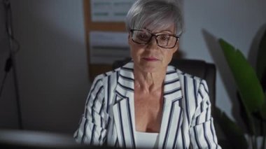 Kısa gri saçlı, gözlüklü, ofis ortamında çalışan, çizgili giyinen yaşlı bir kadın..