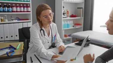 Doktor, bir klinik odasında hastaya danışmanlık yapıyor. Masasında steteskop ve dizüstü bilgisayarı olan iki kadın var..