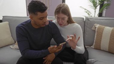 Kadın ve erkek rahat bir oturma odasında kanepede birlikte oturuyor, akıllı telefona odaklanmış, evdeki bağlantıyı ve rahatlamayı sembolize ediyor..
