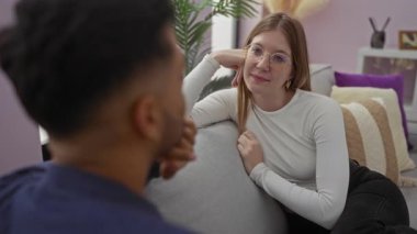 Kadın ve erkek rahat bir oturma odasında kanepede konuşuyor, sıcak bir ilişki ve rahat bir ev ortamı gösteriyorlar..