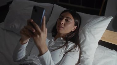 Beyaz yastıklı, rahat bir yatak odasında, beyaz pijama giyen, geceleri evde rahat ve gülümseyen İspanyol bir kadın.