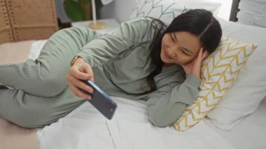 Kadın telefonuyla video görüşmesi yaparken yatakta dinleniyor, yumuşak yastıklı rahat bir yatak odası ayarını vurguluyor, rahatlatıcı bir ev atmosferi yaratıyor..