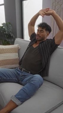Evdeki rahat bir kanepede dinlenen, rahat bir kıyafet giyen, modern dekorlu ve doğal ışık filtresiyle rahat bir oturma odasında eğlenen genç bir adam..