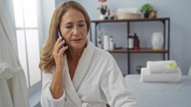 Spa merkezinde orta yaşlı bir kadın bornoz giyiyor, telefonla konuşuyor ve arka planda spa gereksinimleri olan sakin bir sağlık ortamında bir belge okuyor..
