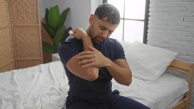 Yatak odasında, gündelik giysiler içinde oturan, dirseğini tutarken acı çeken, arka planda beyaz çarşaflar ve kapalı alanda bitkiler olan genç İspanyol bir adam..