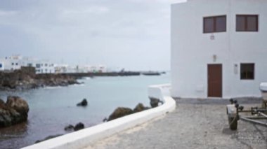 Lanzarote 'de bulanık bir kıyı manzarası, beyaz binalar, okyanus, kayalık kıyılar, bulutlu gökyüzü ve kumlu bir yolda sönmüş bir karavan.