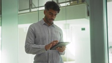 Tabletli genç adam modern ofiste cam duvarlarla duruyor. Odaklanmış ifade ve ev ortamında şık bir görünüm sergiliyor.