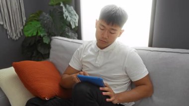 Oturma odasındaki koltukta yeşil bitkiler ve turuncu yastıklı tablet kullanan genç adam rahat ve teknolojik bir ortam yaratıyor..