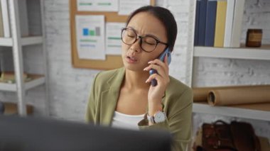 Ofiste telefonla konuşan Asyalı kadın yoğun bir çalışma ortamı sergileyen kitaplar ve çizelgelerle çevrili.