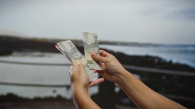 Deniz kıyısındaki bir plajda Endonezya rupia banknotları tutan eller, açık hava ortamında para alışverişini vurguluyor, seyahat ve finansmanı sembolize eden sakin bir deniz zeminine bakılıyor..