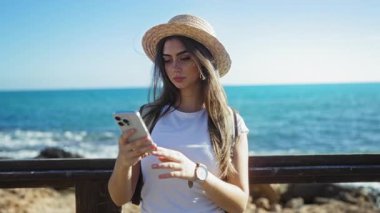 Güneşli bir günde deniz kenarında selfie çeken bir kadın şapka takıyor ve arka planda okyanus olan akıllı bir telefon tutuyor.