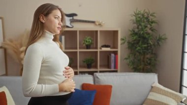 Oturma odasında hamile bir kadın. Arka planda raflar ve gitarla karnını tutuyor..