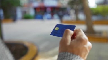 Şehir parkında elinde mavi bir kredi kartı tutan adam, şehir arka planı bulanıklaşırken, açık hava finansal anını yakalıyor..
