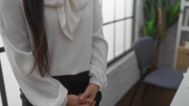 Modern ofiste kolları çapraz duran, zarif beyaz bir bluz giyen bir kadın, kendine güvenini ve profesyonelliğini parlak, havadar bir çalışma alanında gösteriyor..